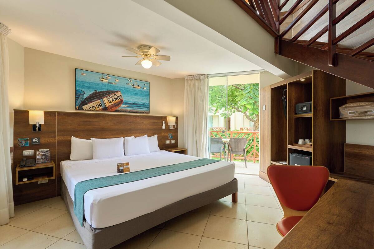Quarto de hotel com cama grande, ventilador de teto, pintura de um barco, escrivaninha e uma cadeira.