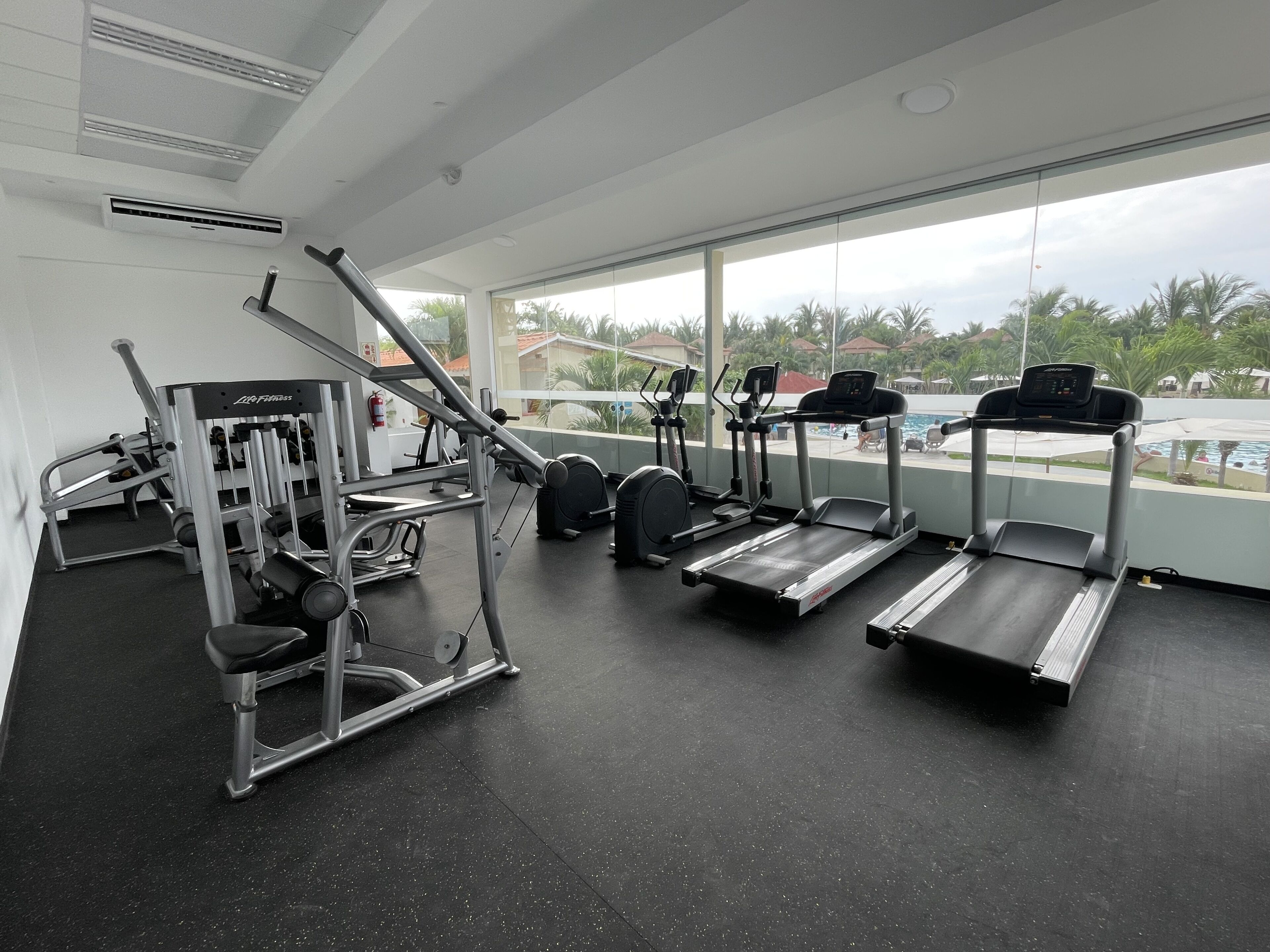 Ett gym med löpband, styrketräningsmaskiner och utsikt över en swimmingpool och palmer.