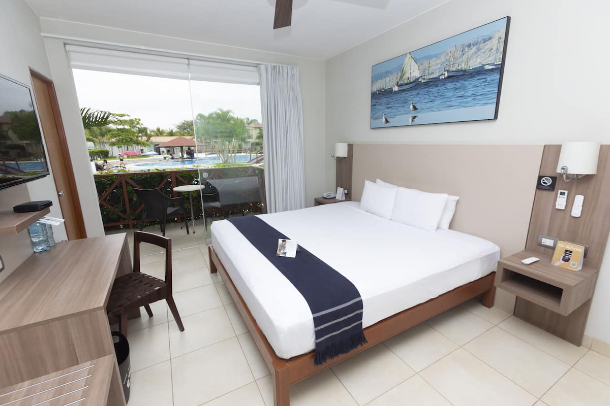Quarto de hotel com cama, escrivaninha, cadeira e vista para uma piscina ao ar livre.