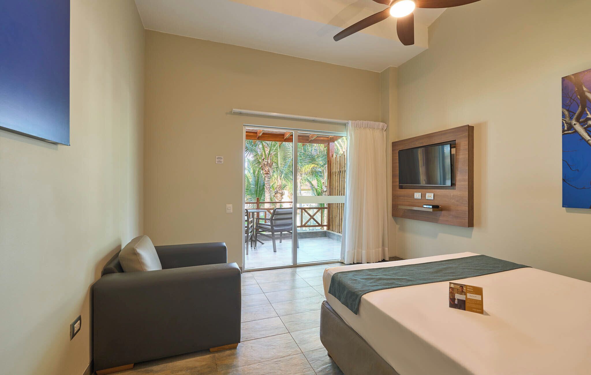 A hotel room with a bed, a TV mounted on the wall, a ceiling fan, and a view of a patio with a table and chairs.