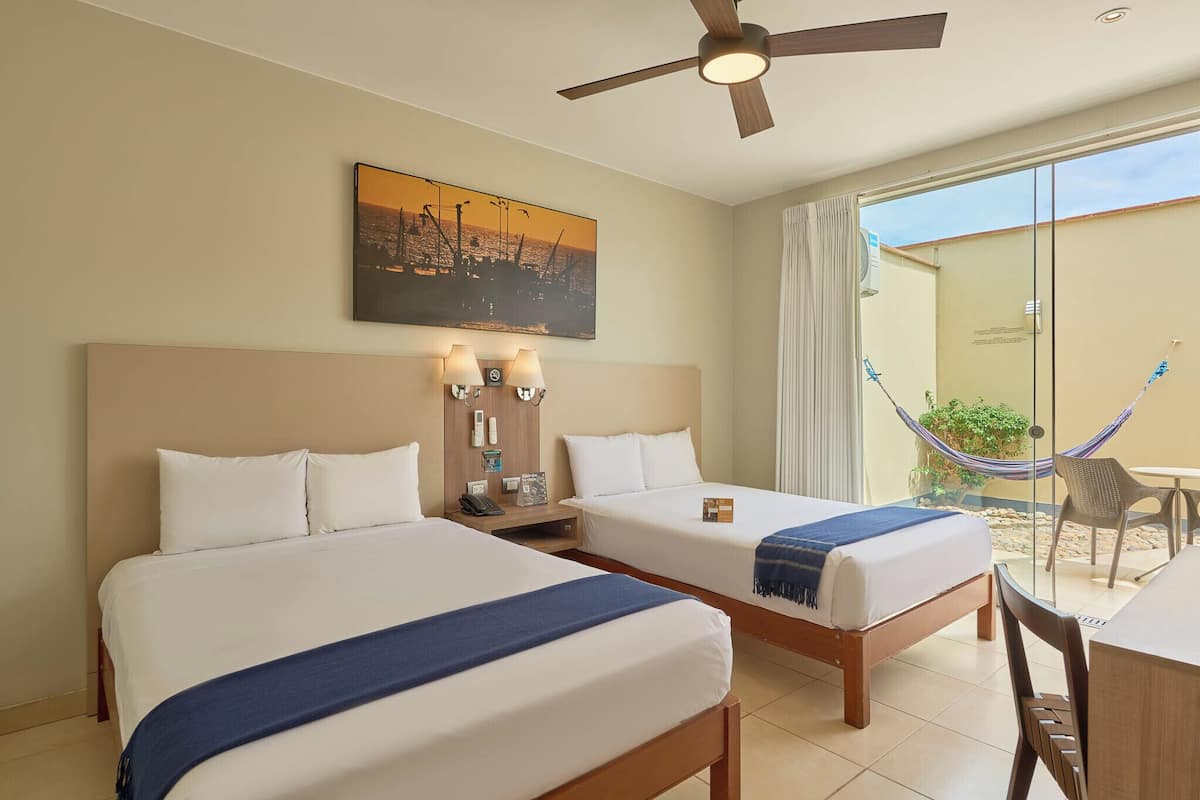 Quarto de hotel com duas camas, um ventilador de teto, mesa de cabeceira com luminárias, um quadro na parede e vista para uma rede e um pátio lá fora.