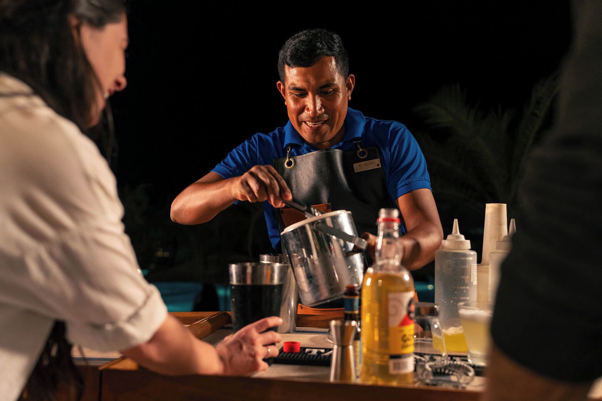 A person pouring liquid from a pot into a glass, with condiments and a bottle on the table.