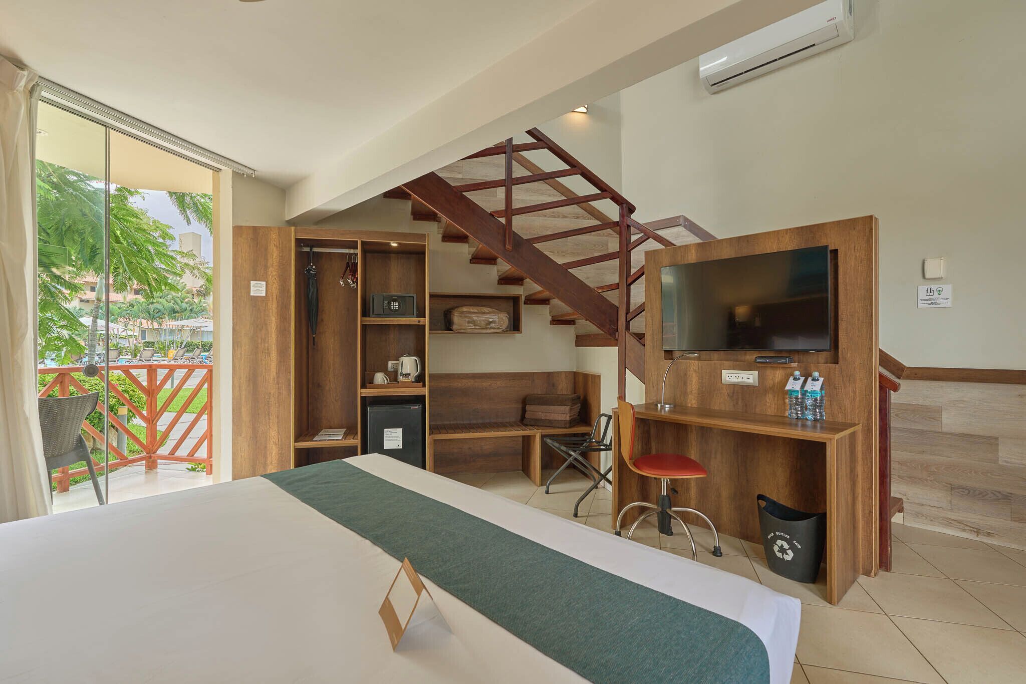 A hotel room with a bed, a TV, a desk, a chair, a bookshelf, a small table, a trash bin, and a staircase.