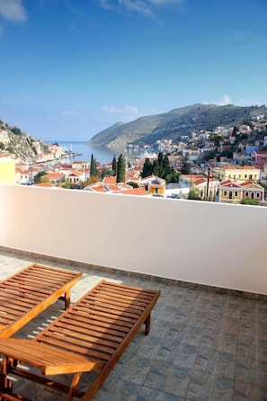 A terrace with wooden loungers overlooking a coastal town with buildings and a mountain in the background. - Eos Studios (Symi)