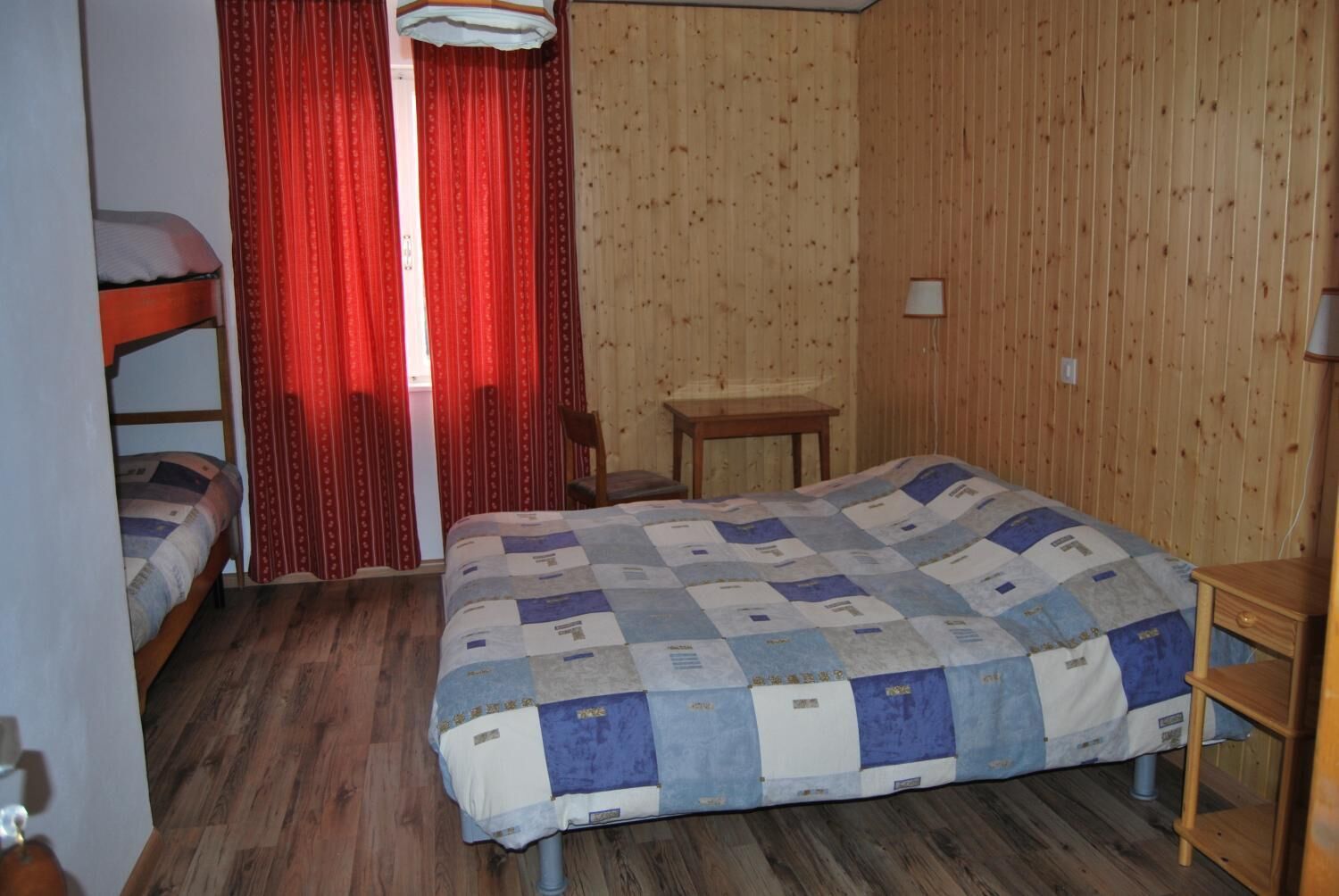 Une pièce avec un plancher en bois, un lit recouvert d’une courtepointe, une chaise, une petite table et une fenêtre ornée de rideaux rouges.