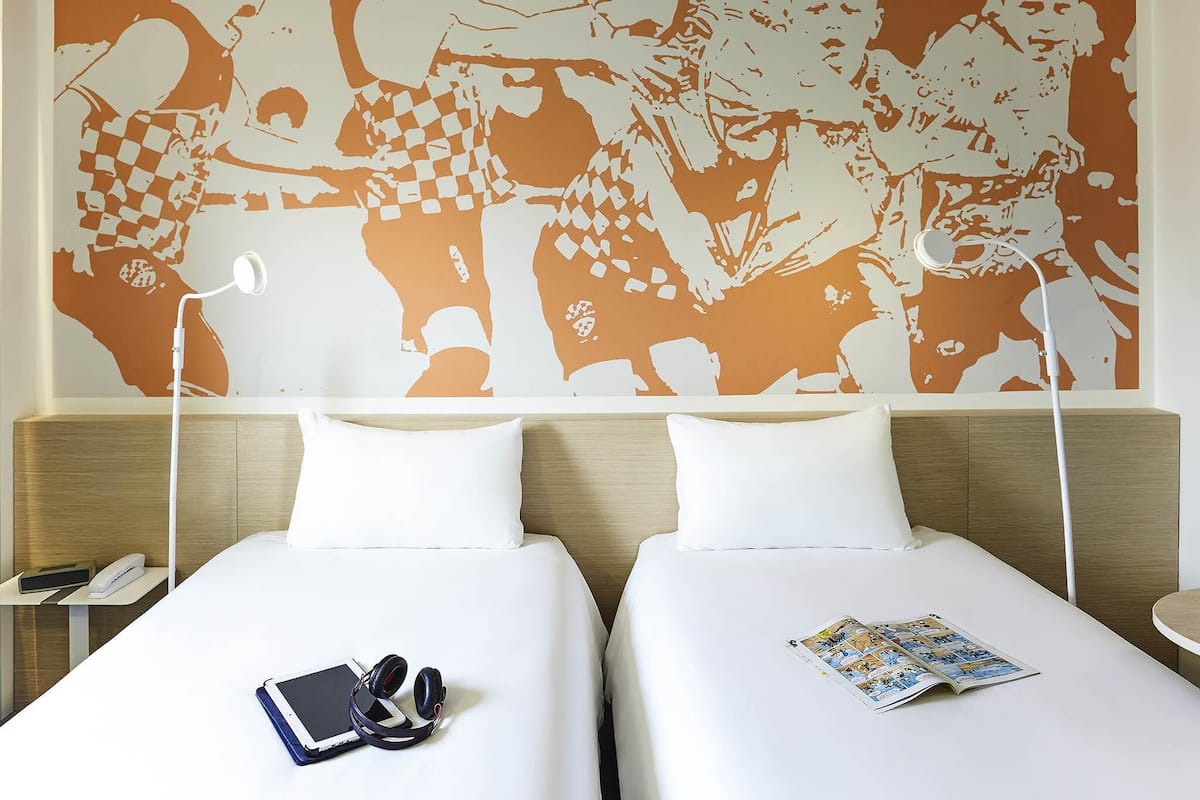 Ein Hotelzimmer mit zwei Betten, einem großen abstrakten Wandbild, einem Nachttisch mit Lampe und einer Zeitschrift auf einem der Betten.