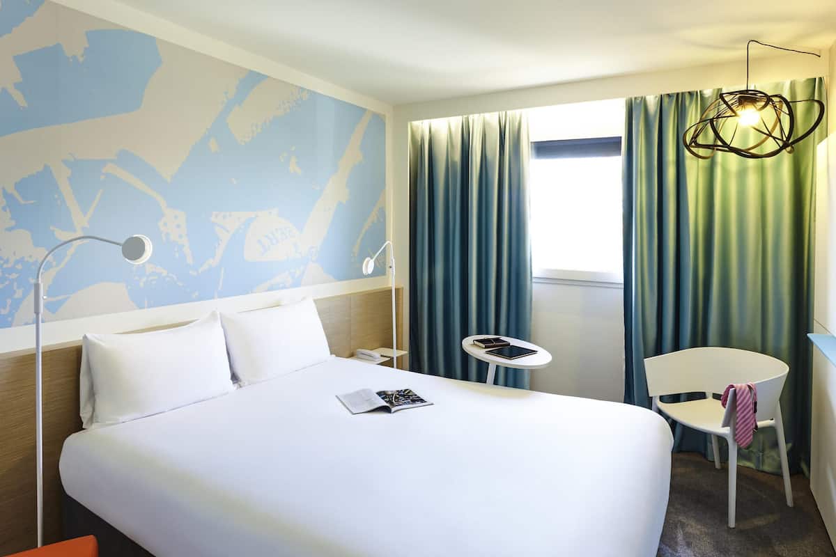 Ein Hotelzimmer mit Bett, Schreibtisch, Stuhl, Lampe und einem Fenster mit Vorhängen.