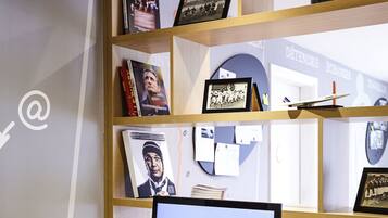 A wooden desk with a computer, keyboard, mouse, and printer. There is a white chair in front of the desk. The wall behind the desk has shelves with books and framed pictures.