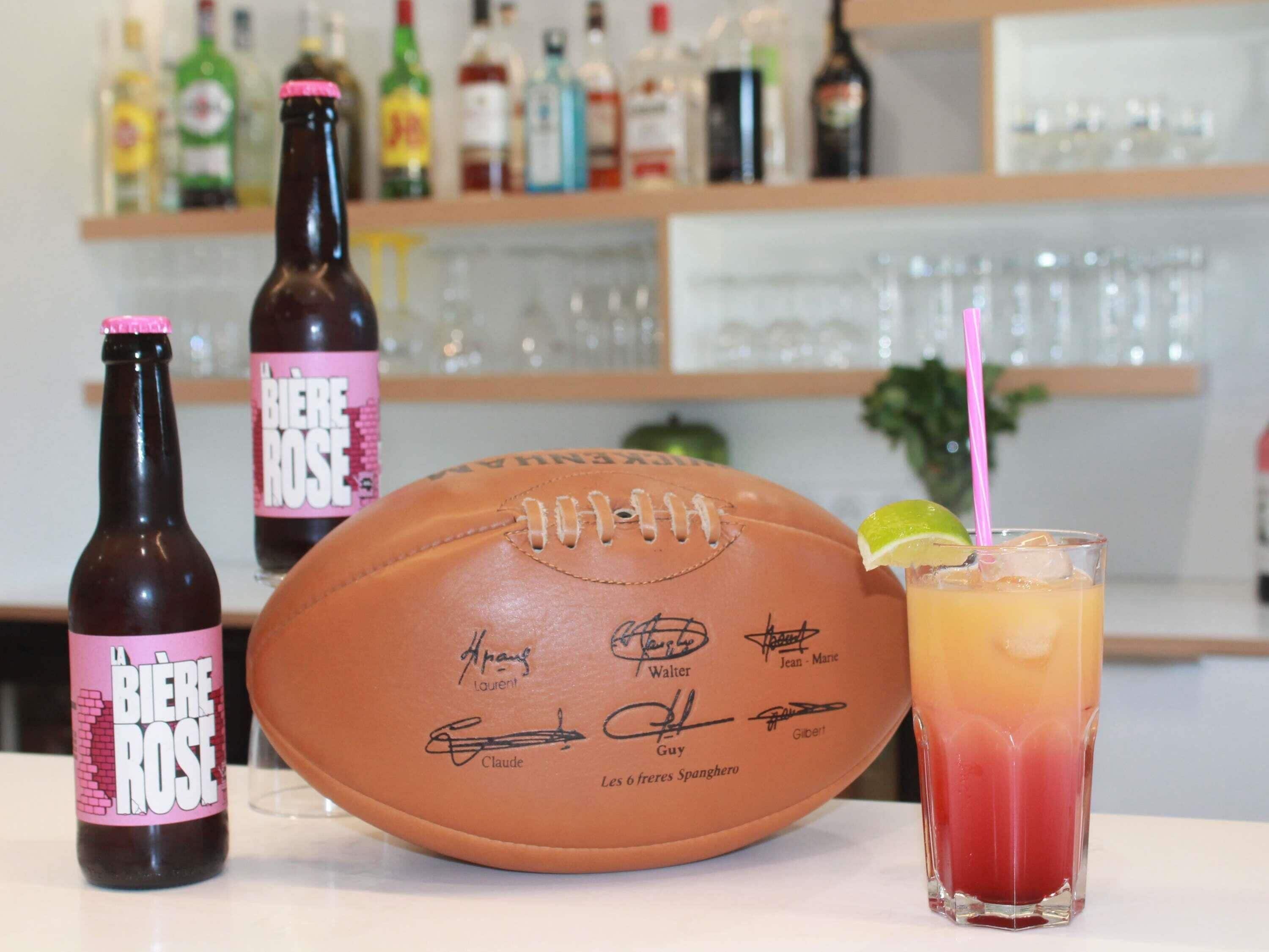 Una pelota de fútbol, una botella de cerveza y una copa de cóctel sobre una mesa con un bar al fondo.
