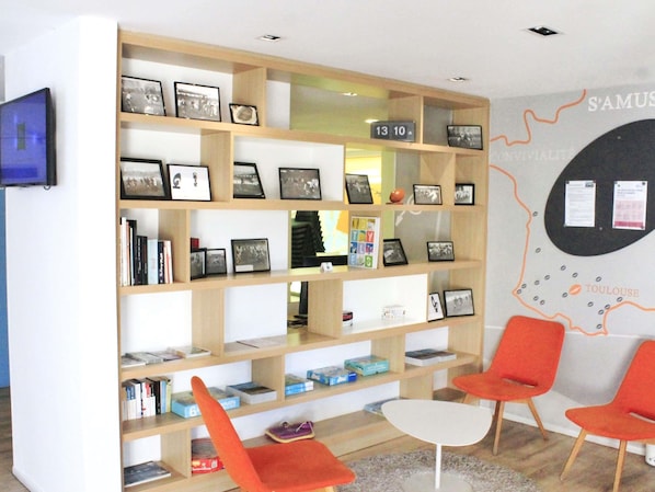 A room with a bookshelf filled with books and framed photos, a white table, and two orange chairs. - ibis Styles Toulouse Nord Sesquières (Toulouse)