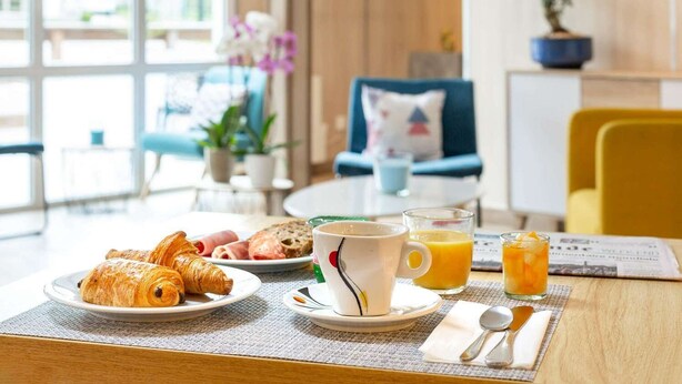 A breakfast table with croissants, eggs, and juice. There is a chair and a potted plant in the background. - Brit Hotel Rouen Barentin (Barentin)