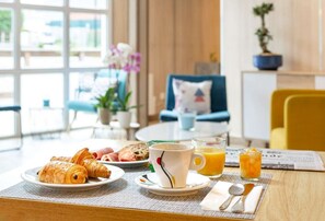 Una mesa de desayuno con croissants, huevos y jugo. Se ve una silla y una planta en maceta al fondo.