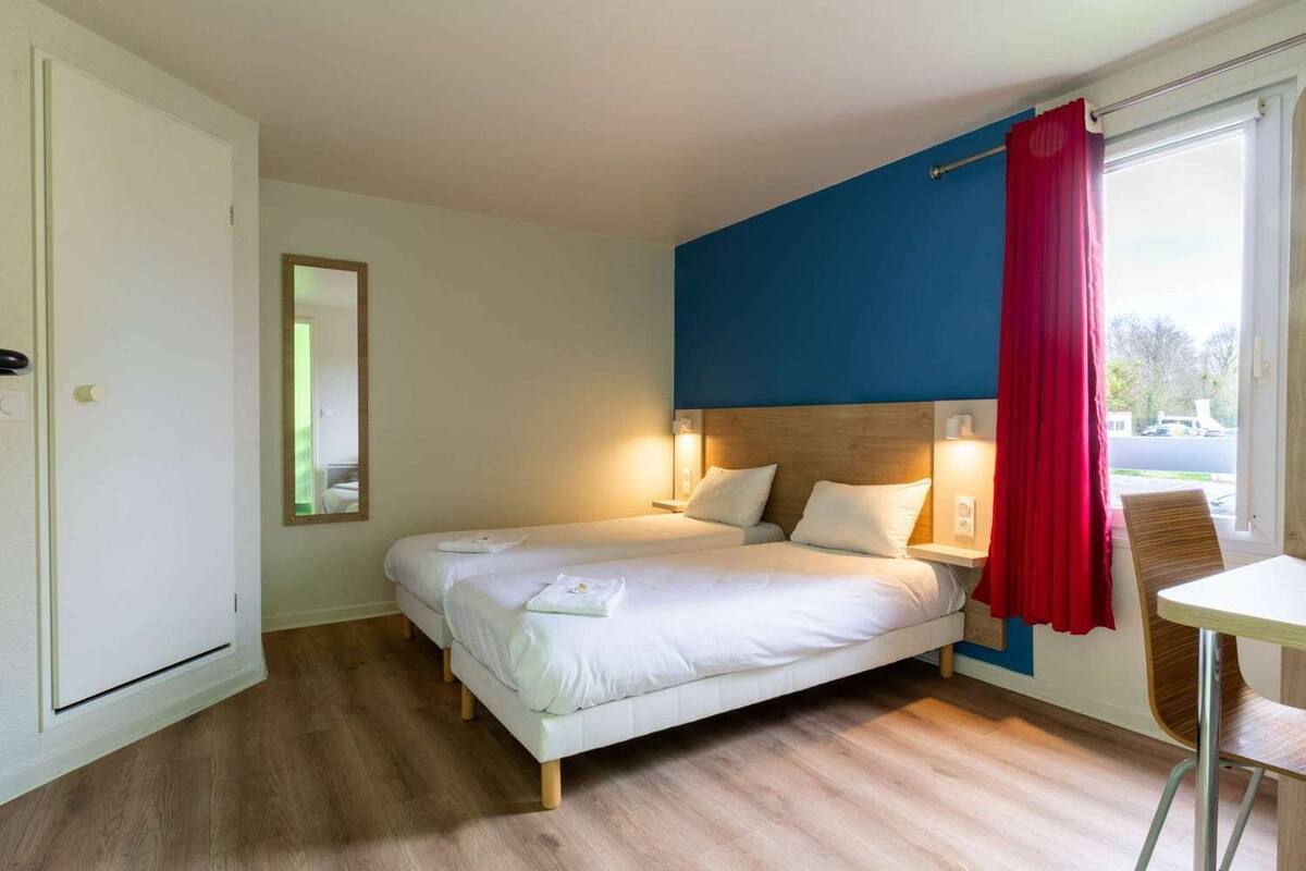 A hotel room with a bed, bedside tables, a mirror, a window with a red curtain, and a view of the outside.