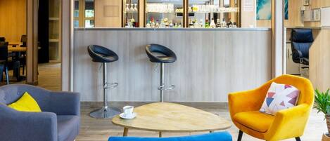 Un hall d’hôtel moderne avec un coin bar, deux tabourets de bar, une table ronde avec une tasse et un fauteuil jaune.