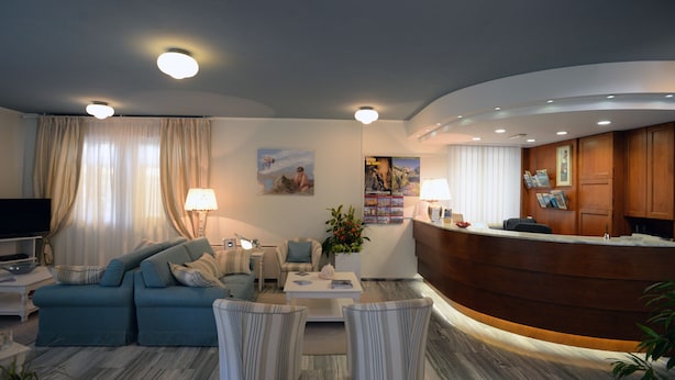 A modern hotel lobby with a reception desk, seating area with sofas and armchairs, a flat-screen TV, and a large window with curtains.