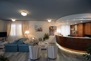 Eine moderne Hotelhalle mit einer Rezeption, einer Sitzecke mit Sofas und Sesseln, einem Flachbildfernseher und einem großen Fenster mit Vorhängen.