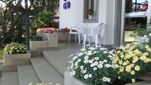 A building with a concrete staircase leading to a white door, a small outdoor seating area with white chairs, and flower planters with yellow and white flowers.