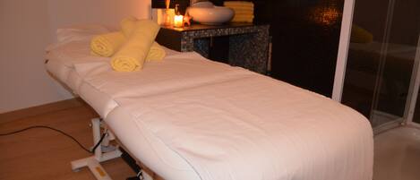 A single bed with a white mattress and pillows, a small table with a candle and towels, and a shower area with a glass door.