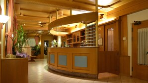 A hotel lobby with a reception desk, wooden paneling, and a tiled floor. - Hotel Ladina (Campitello di Fassa)