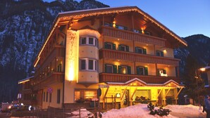 A multi-story hotel with a balcony, illuminated by warm lights, set against a snowy mountain backdrop. - Hotel Ladina (Campitello di Fassa)