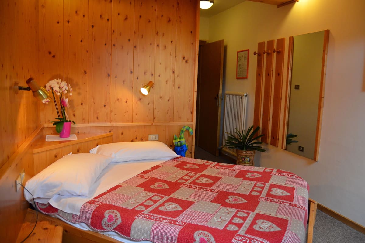 A wooden-paneled bedroom with a bed, a nightstand with a vase of flowers, a mirror, and a radiator.
