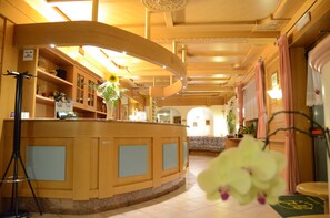 A reception area with a wooden counter, shelves with various items, and a floral arrangement. - Hotel Ladina (Campitello di Fassa)
