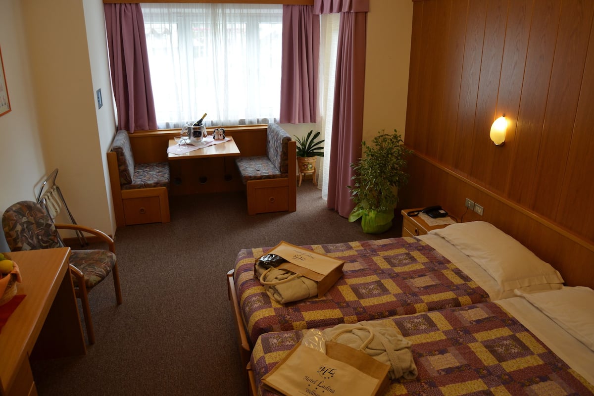 A hotel room with a bed, a desk, a chair, a small table, and a potted plant.
