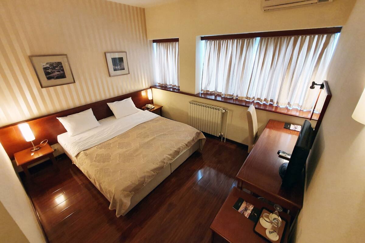 Quarto de hotel com uma cama grande, mesas de cabeceira, escrivaninha, cadeira, radiador e janela com cortinas.