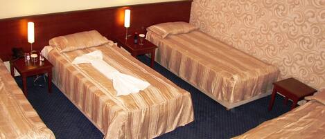 A hotel room with four beds, each with a wooden headboard and a patterned bedspread. There are bedside tables with lamps and a telephone on one side.
