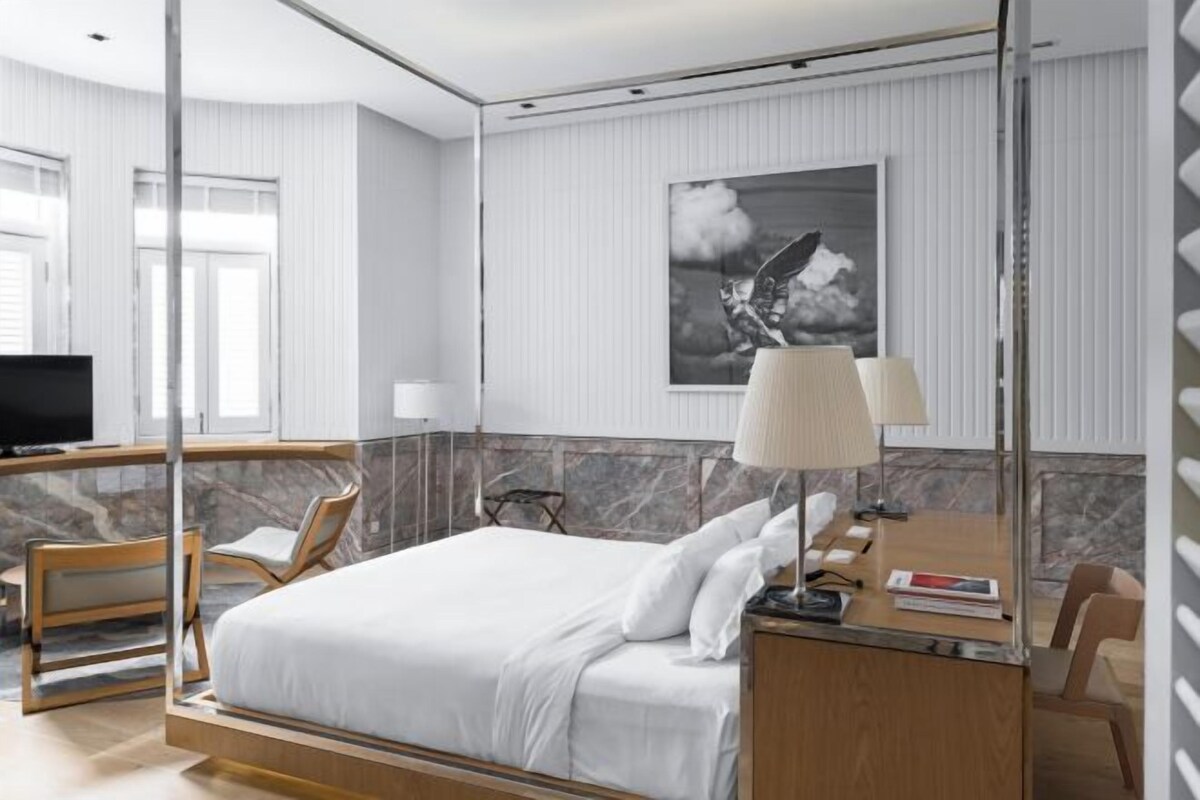 Ett hotellrum med en säng, ett skrivbord med en lampa, en stol, en tv och en inramad tavla på väggen.