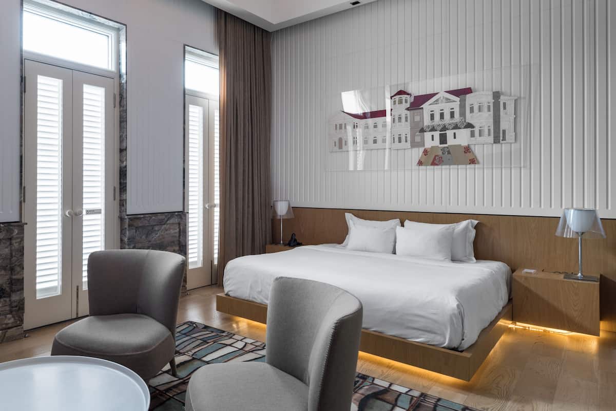 A hotel room with a large bed, two armchairs, a small table, a decorative wall piece, and a view of the outside through the window.