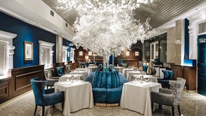 A restaurant with a central seating area featuring a large, white, tree-like structure. Tables are arranged around this structure, with blue upholstered chairs. The room has wooden paneling on the walls and a herringbone-patterned floor. - Macalister Mansion (George Town)