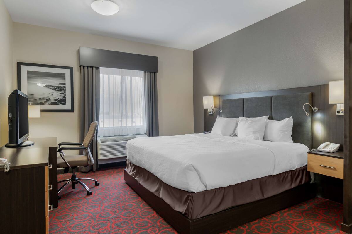 Habitación de hotel con una cama grande, un escritorio con computadora, una silla, una ventana con cortinas y un cuadro en la pared.
