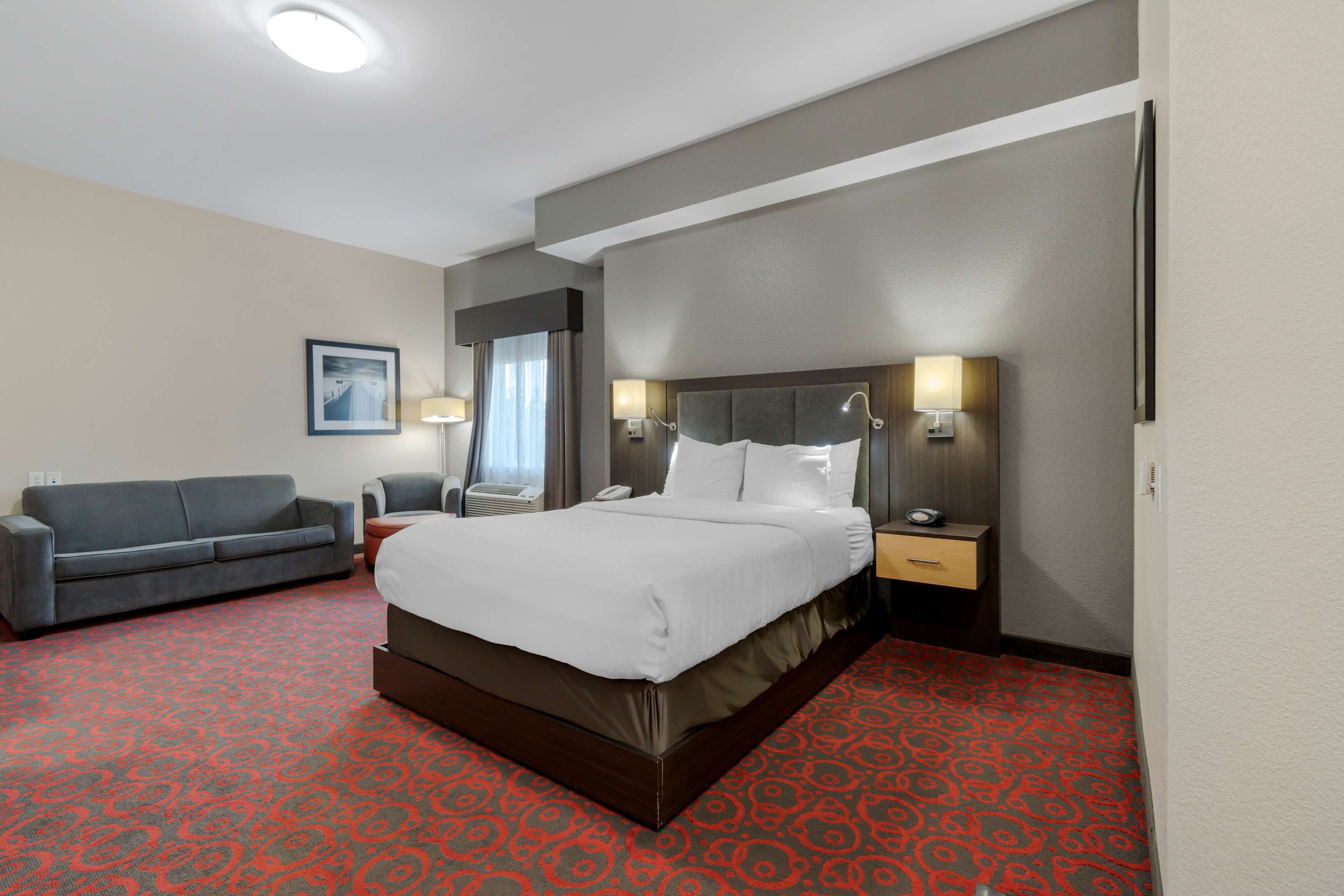 Ein Hotelzimmer mit einem großen Bett, einem grauen Sofa, einem Schreibtisch mit einer Lampe und einem Bild an der Wand.