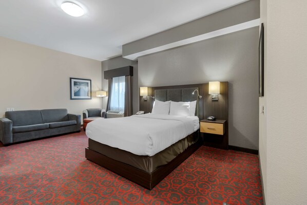 A hotel room with a large bed, a grey sofa, a desk with a lamp, and a picture on the wall.