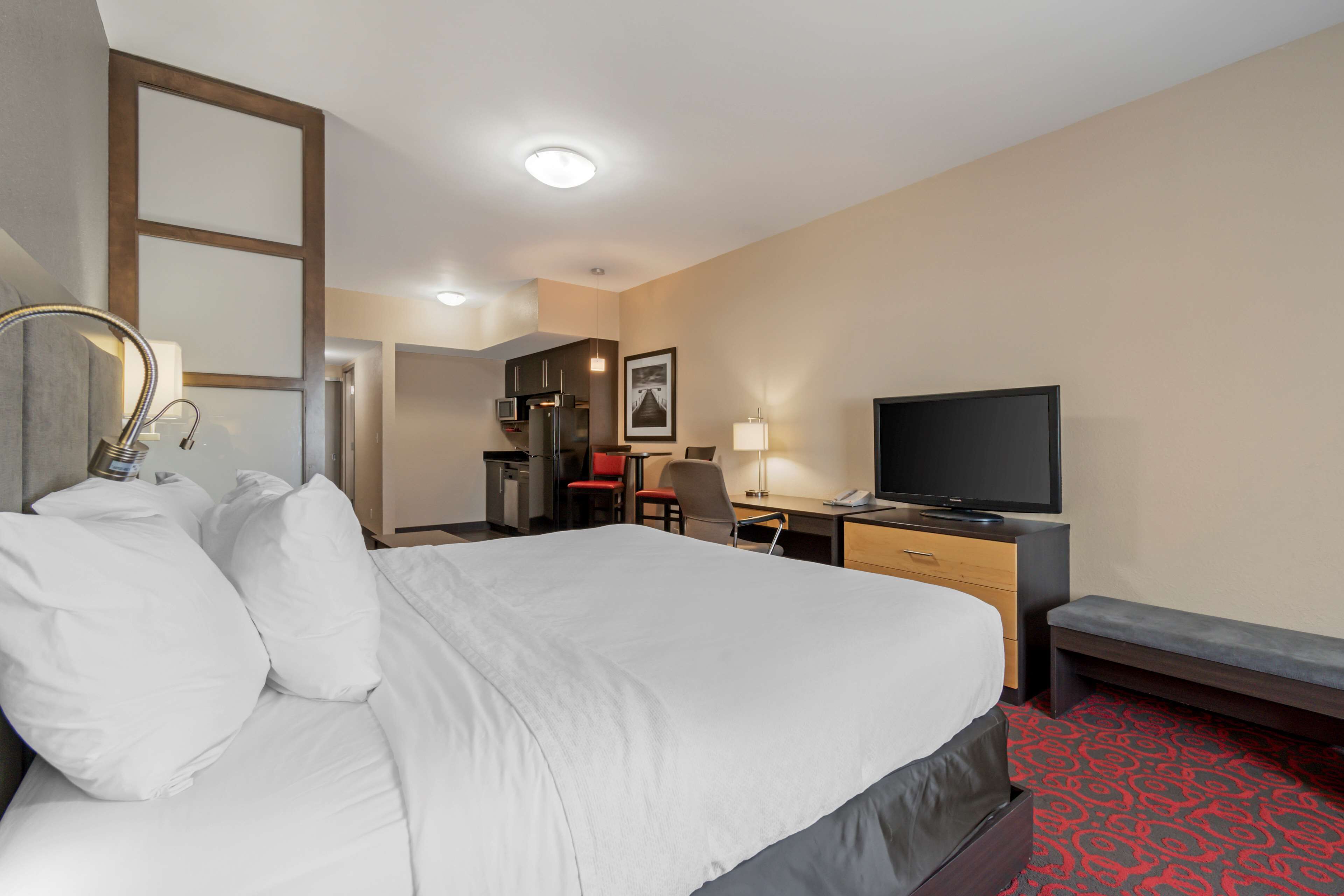 A hotel room with a large bed, a desk with a computer, a television, and a kitchenette in the background.