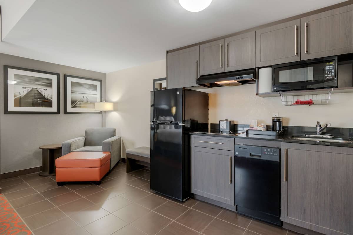 Una cocina moderna con armarios grises, un refrigerador negro, lavavajillas y un microondas. Hay un sillón gris y una pequeña mesa con una lámpara.