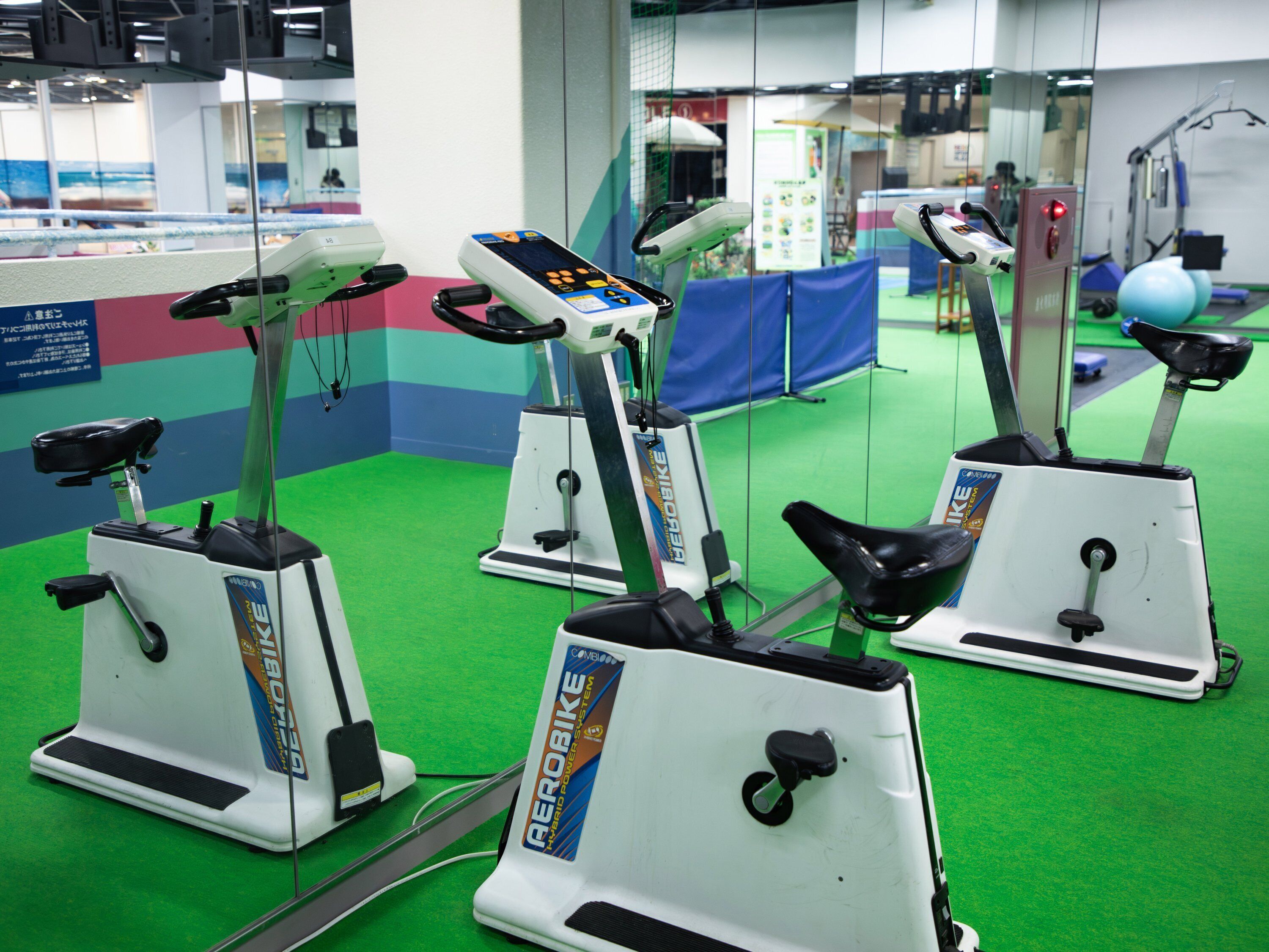 Three stationary bikes in a gym with green flooring and a blue and red wall.
