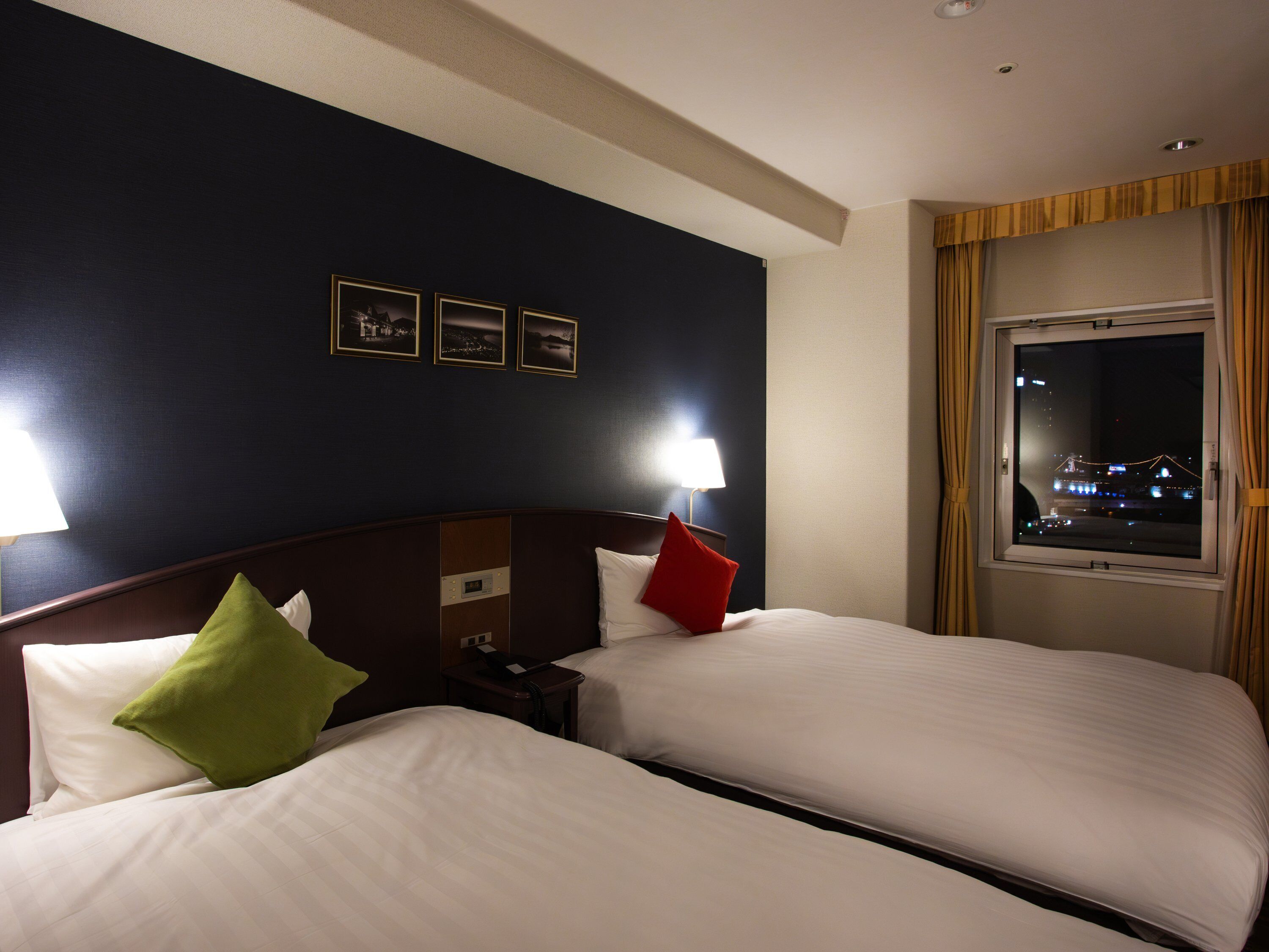 A hotel room with two beds, a nightstand, a lamp, a window with a view of a cityscape, and three framed pictures on the wall.