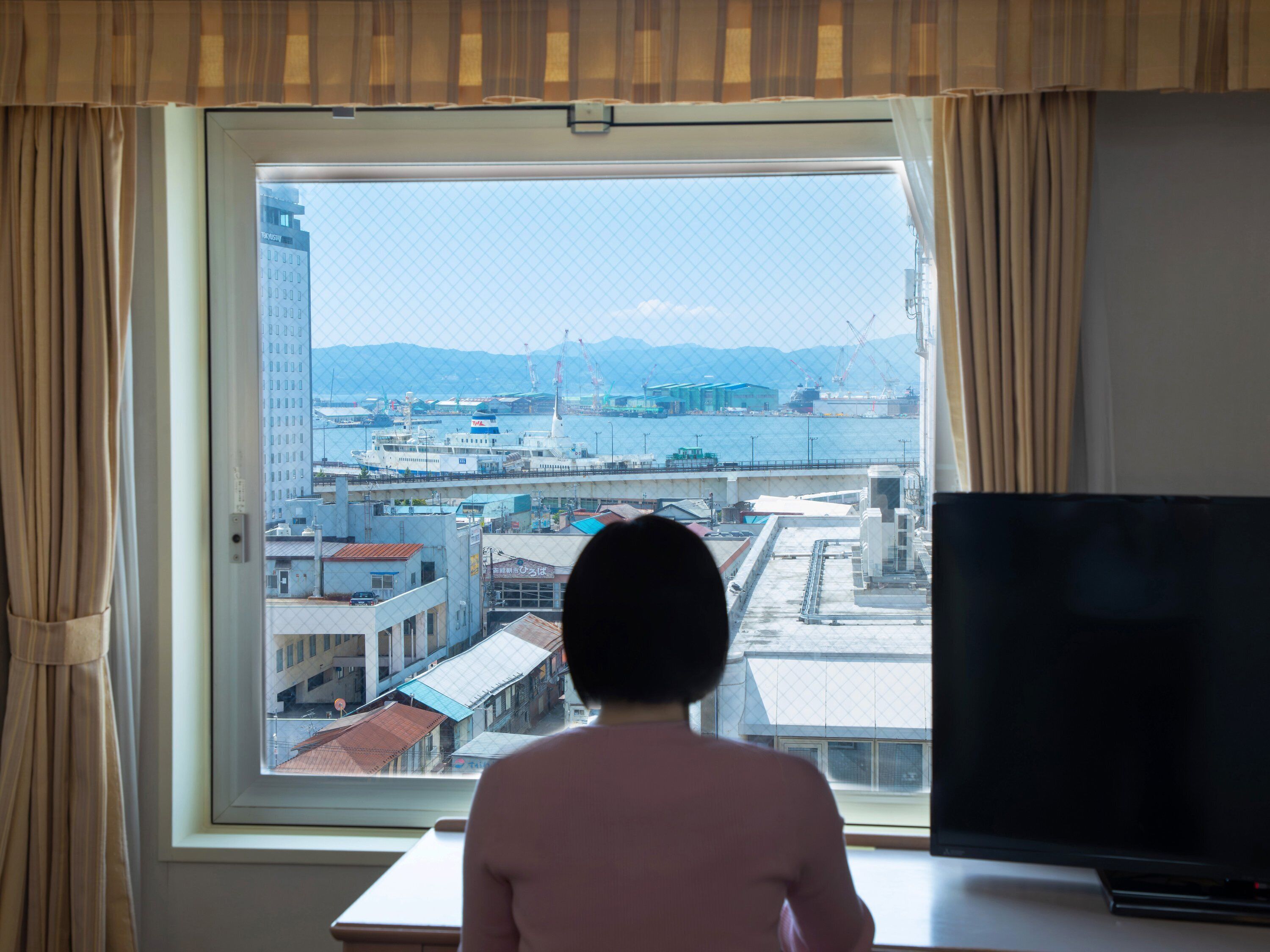 A room with a large window overlooking a harbor, a television on a table, and a person sitting at the window.