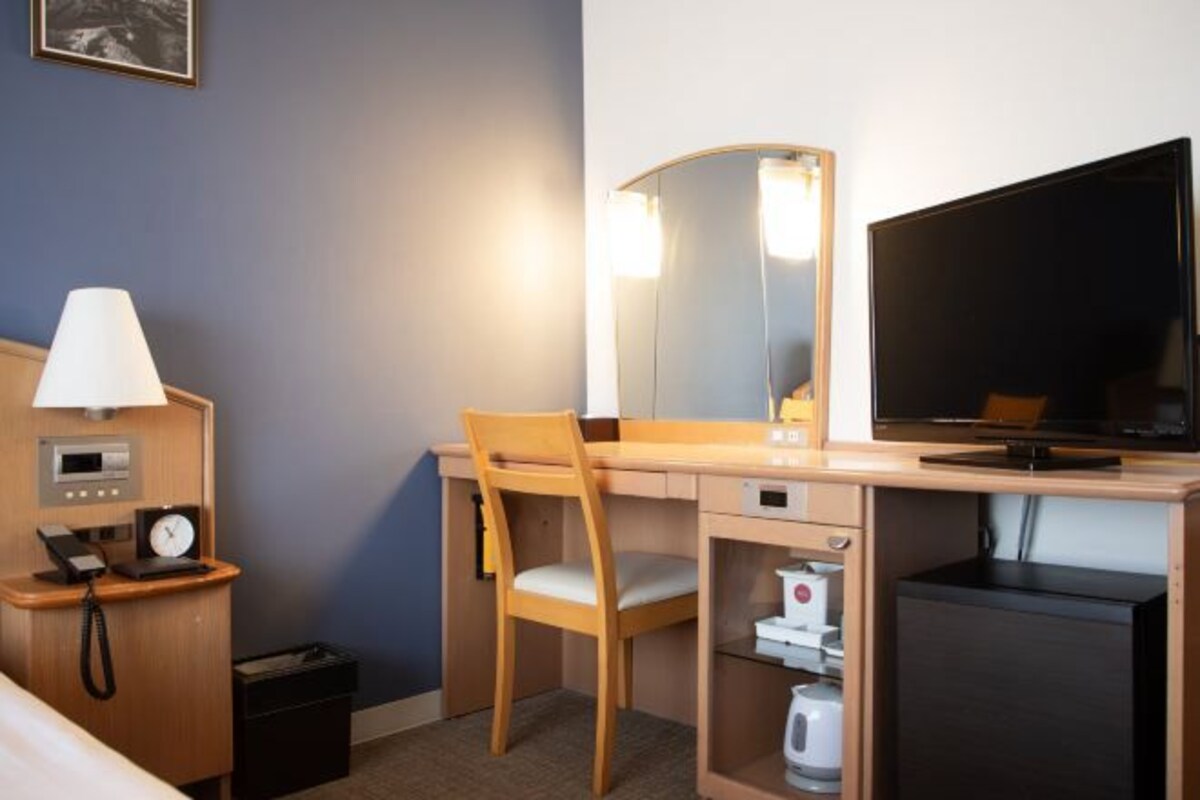 A hotel room with a bed, a desk with a chair, a television, a mirror, a lamp, a clock, and a mini-fridge.