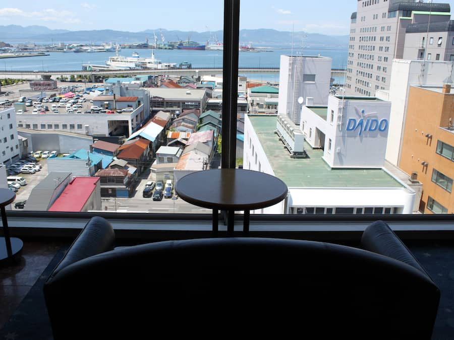 Vista da janela com uma mesa e cadeiras, com vista para um porto com navios e edifícios.