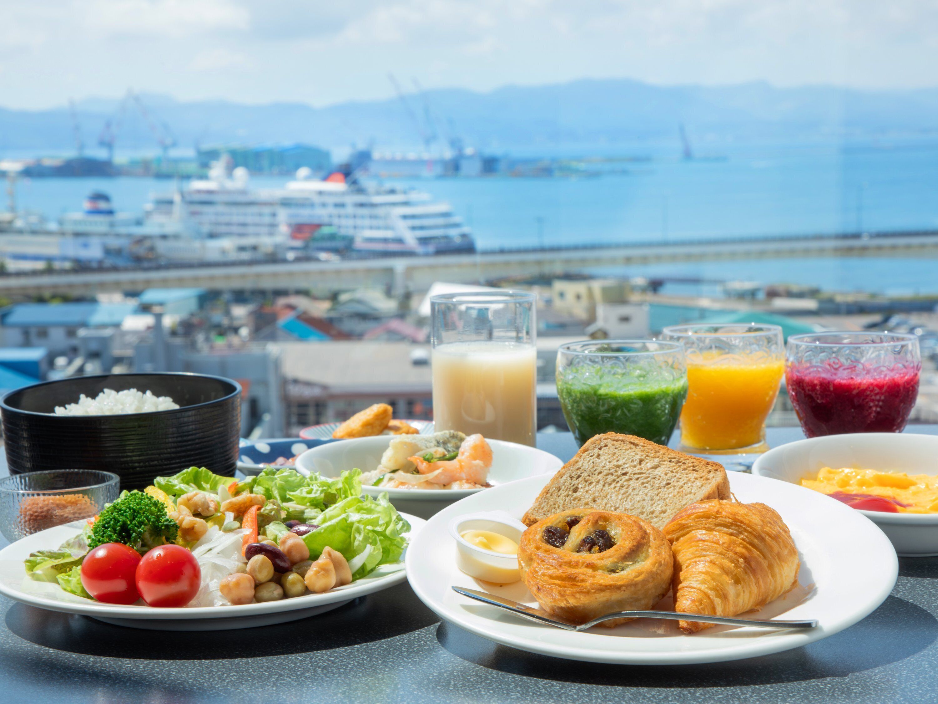 Une table dressée avec divers éléments pour le petit-déjeuner, notamment des toasts, des croissants et une salade, et en toile de fond, une vue sur un port et des navires.