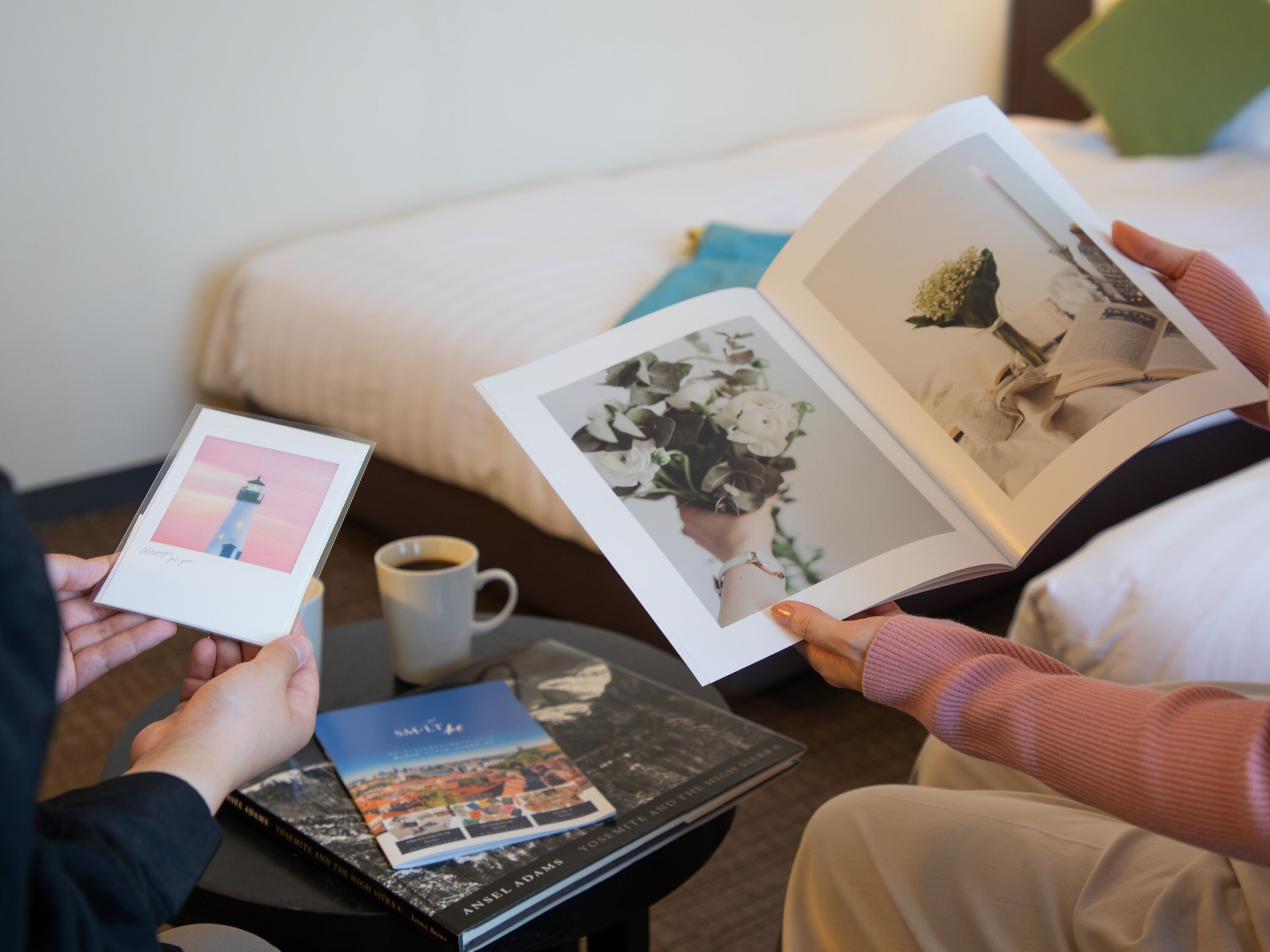 A person holding a book with a photograph of a lighthouse, another book with a cityscape cover, and a cup of coffee on a table.
