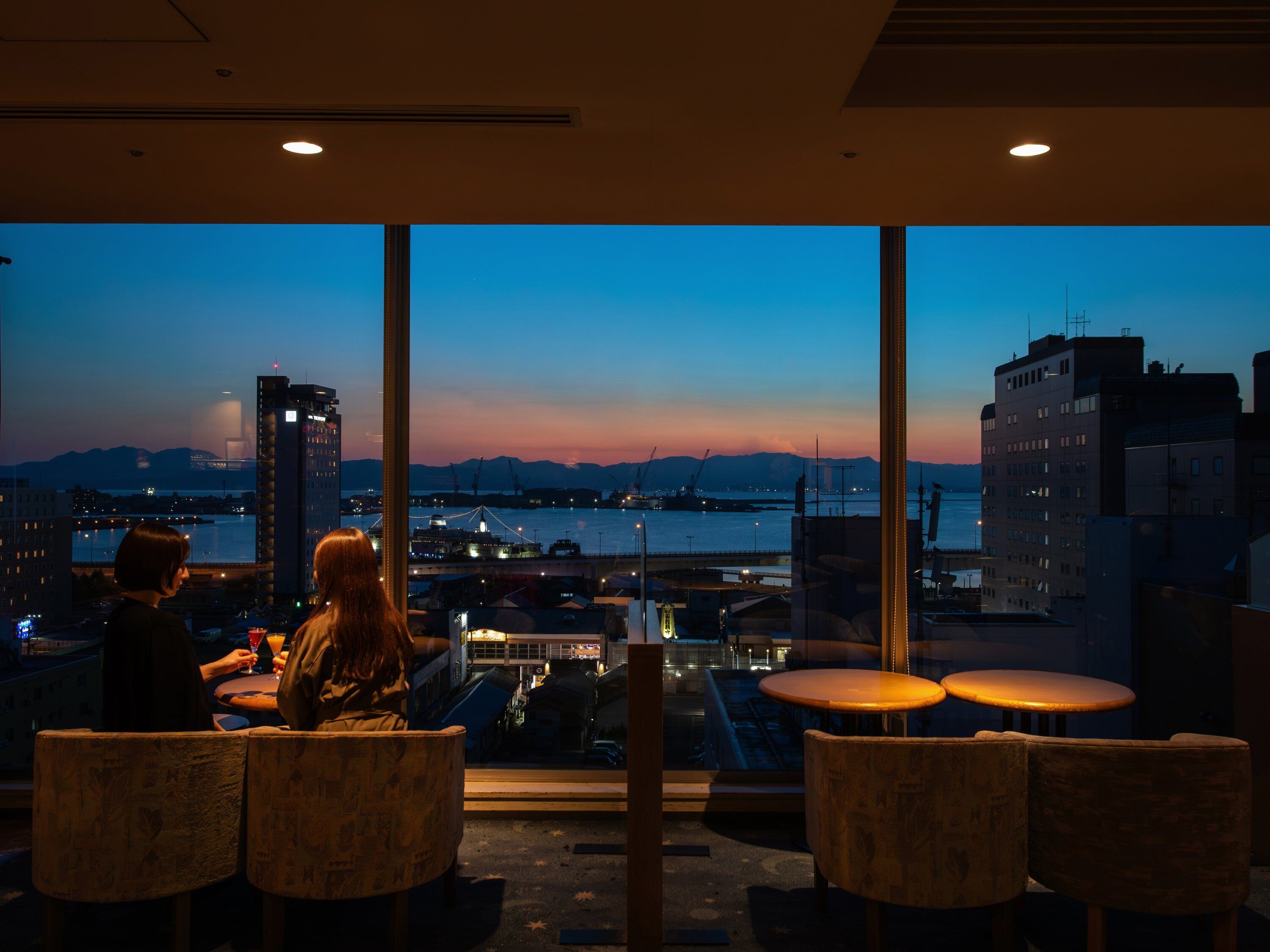 Un restaurante con amplios ventanales que ofrece vistas a un paisaje urbano y al agua al atardecer.