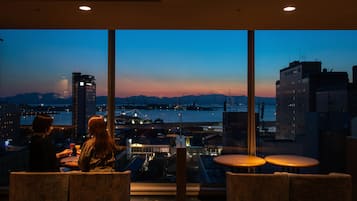 Un restaurante con amplios ventanales que ofrece vistas a un paisaje urbano y al agua al atardecer.