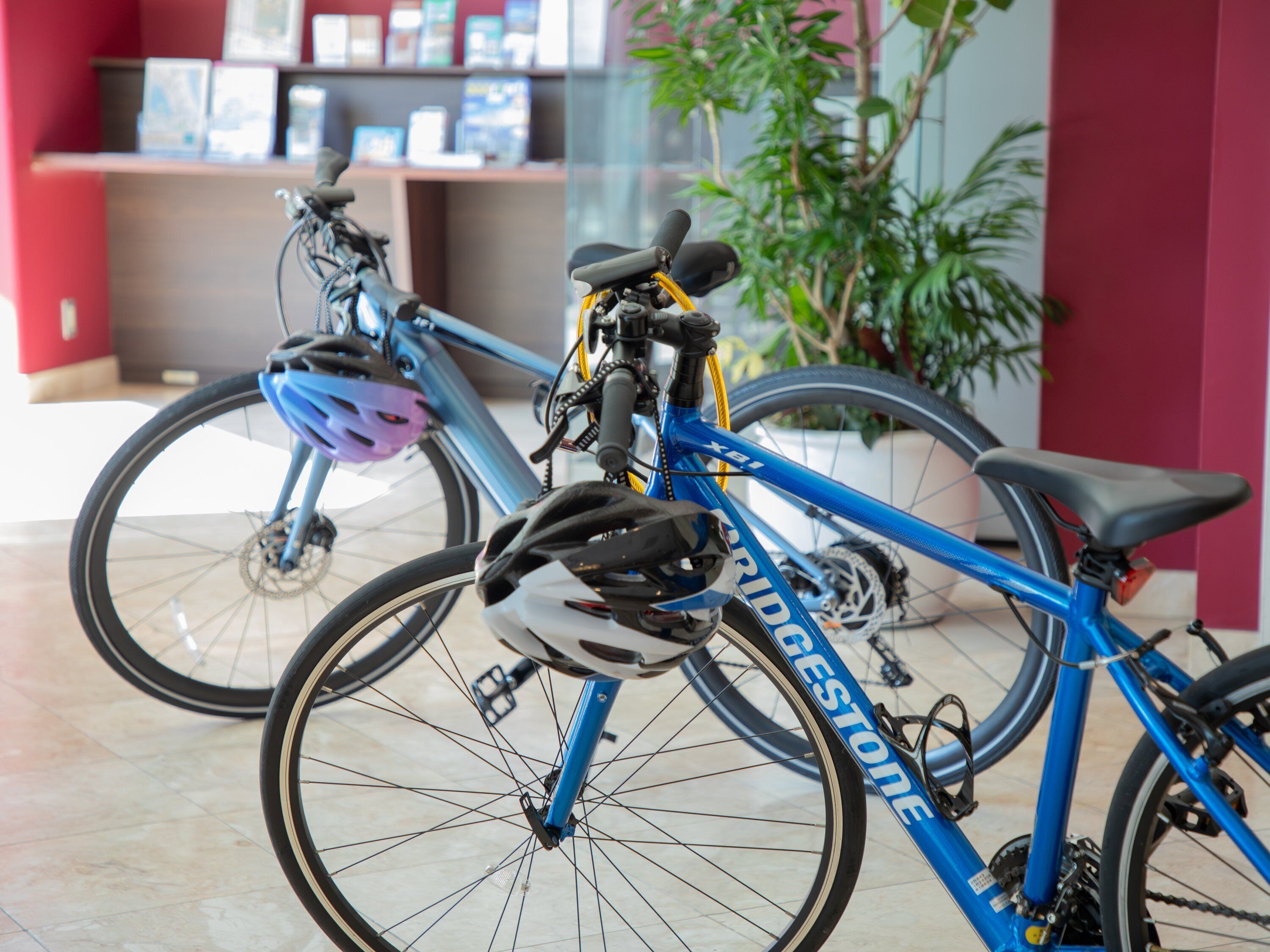 A blue bicycle with a helmet and lock, parked indoors with a plant and a shelf in the background.