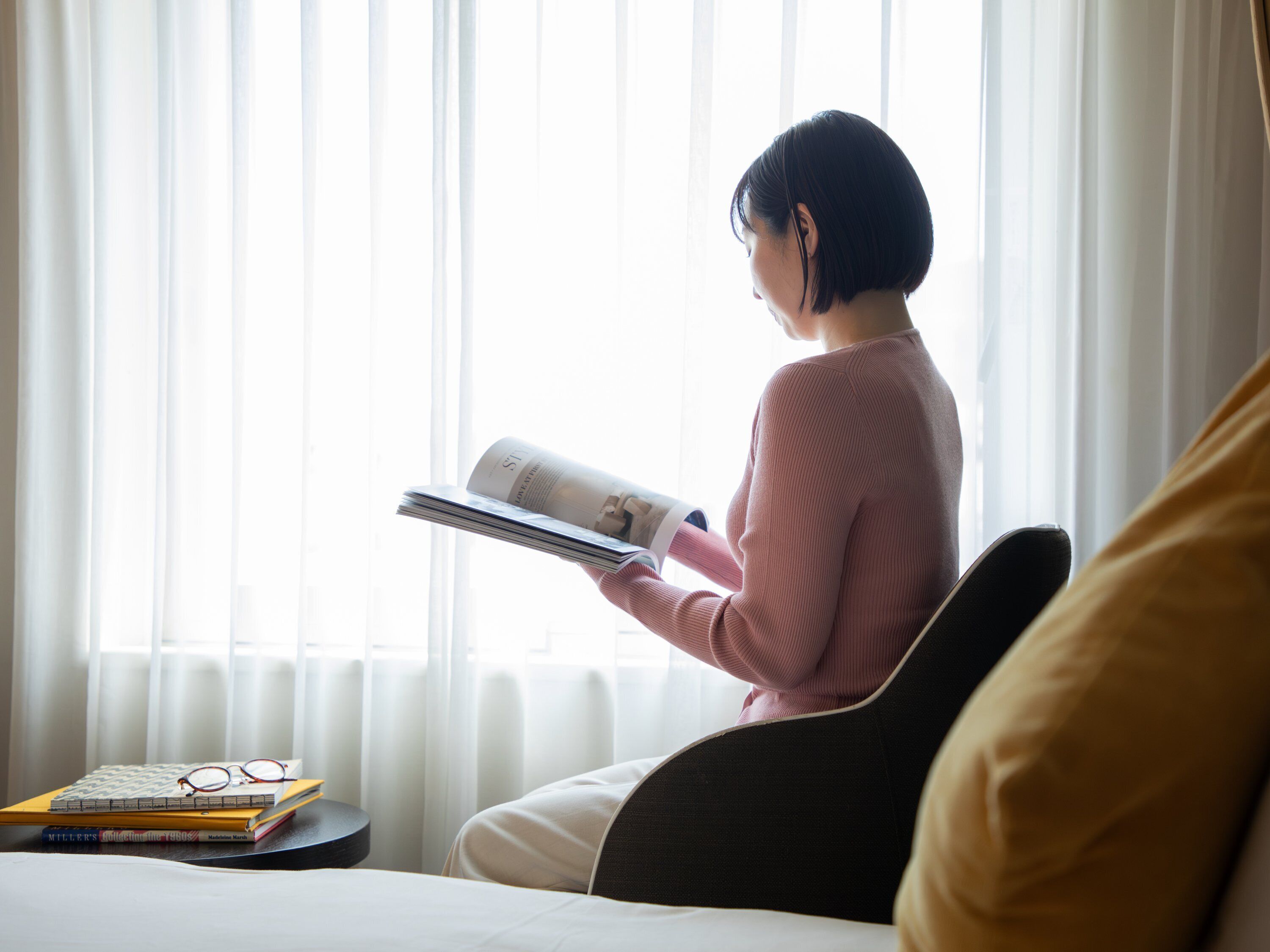 Une personne est assise près d’une fenêtre, des rideaux sont tirés, et elle est en train de lire un magazine. On aperçoit un lit recouvert d’une couverture blanche, une chaise noire et une petite table sur laquelle se trouvent des livres et des lunettes.