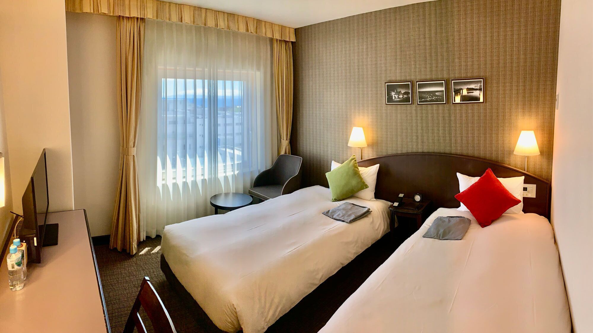 A hotel room with two beds, a desk, a chair, and a view of the cityscape through the window.