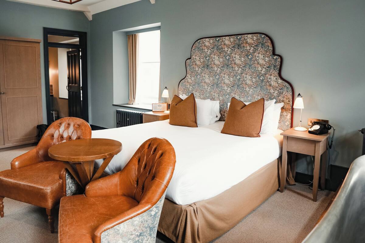 Une chambre d’hôtel avec un grand lit, deux fauteuils en cuir marron, une petite table ronde, une table de chevet en bois avec une lampe, et une grande tête de lit à motifs floraux.